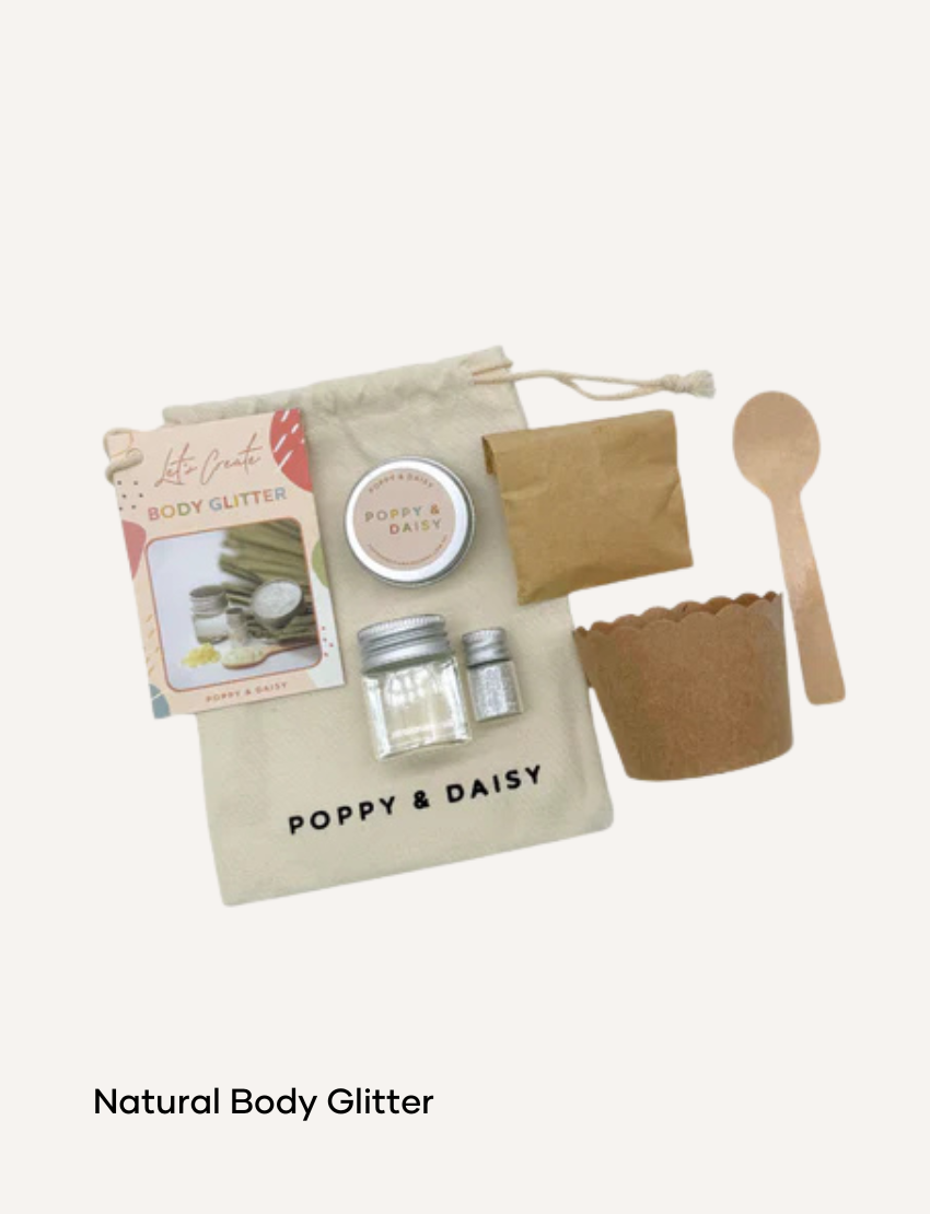 A hand holds a small tin of Lavender Lip Balm. Below it, a cloth pouch labeled "Poppy & Daisy" contains a glass vial, a small wooden stick, and a card with floral illustrations. The background reveals a knitted blanket.