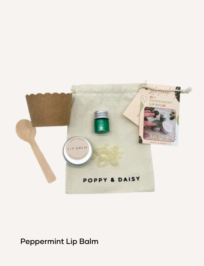 A hand holds a small tin of Lavender Lip Balm. Below it, a cloth pouch labeled "Poppy & Daisy" contains a glass vial, a small wooden stick, and a card with floral illustrations. The background reveals a knitted blanket.