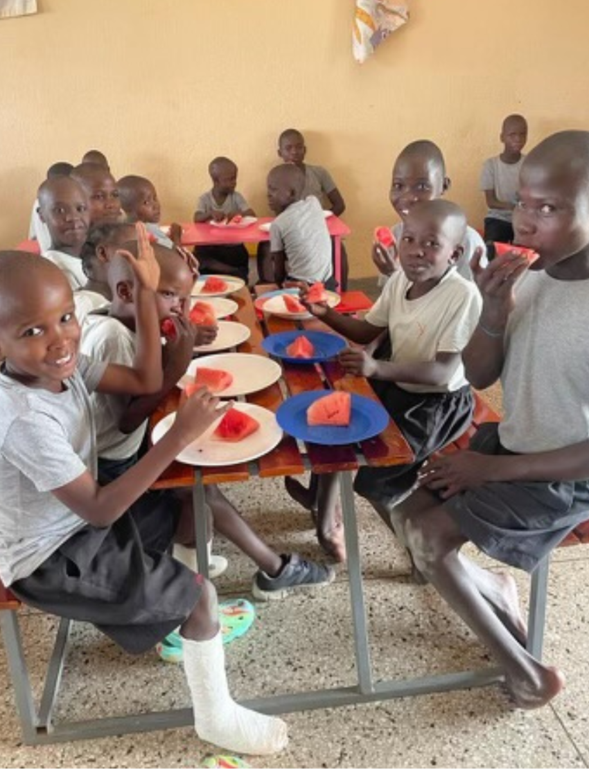 Children in a classroom setting enjoy slices of watermelon during a mealtime, highlighting a charity initiative.