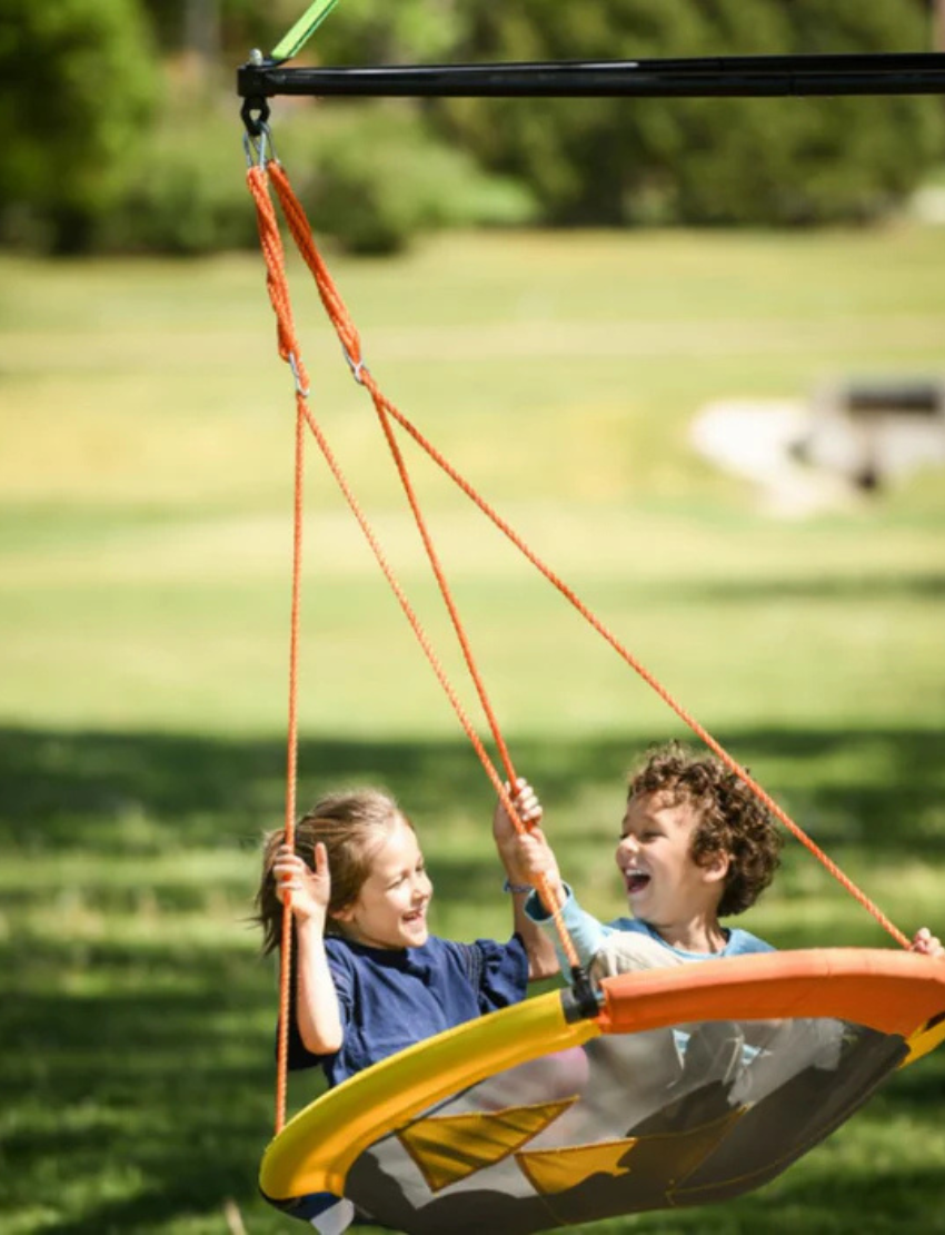 A circular platform swing with yellow, blue, and orange sections hangs by ropes. The product box shows kids using the b4Adventure Sky Adventure Swing Chair with the text "Adventure Sky Swing," inviting imaginative adventures for all ages.